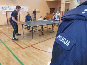 sala gimnastyczna, stół do tenisa, policjant, uczniowie, na pierwszym planie ręka w mundurze z napisem policja