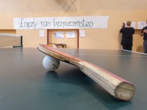 sala gimnastyczna, stół do tenisa, paletka tenisowa i piłeczka, w tle napis łączy nas bezpieczeństwo