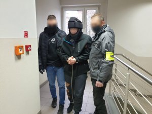 policjanci prowadzą zatrzymanego w kajdankach i hełmie ochronnym