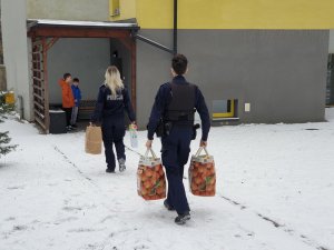 policjant, policjantka idą do budynku z torbami wypełnionymi słodkościami i artykułami spożywczymi