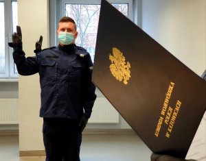 Zdjęcie kolorowe - sala odpraw, policjant stoi w postawie zasadniczej w trakcie ślubowania, na pierwszym planie teczka z orłem i napisem Komenda Wojewódzka Policji w Katowicach