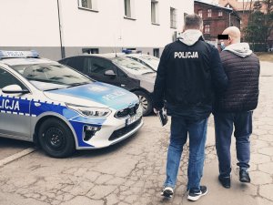 parking komendy, stojące radiowozy po lewej, na pierwszym planie policjant prowadzi zatrzymanego