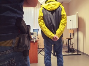 policjant z bronią przy pasie, w tle zatrzymany 25-latek w pomieszczeniu do wykonywania zdjęć sygnalitycznych