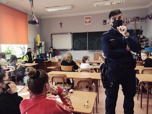 zdjęcie z wizyty policjantów w szkole - policjant dzielnicowy rozmawia z dziećmi siedzącymi w ławkach szkolnych, w tle policjant drogówki i nauczycielka