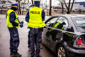 trzech policjantów przy czarnym samochodzie w trakcie kontroli, w tle ulica i budynek magazynowy
