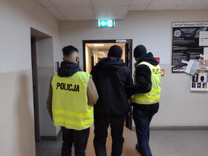 dwóch policjantów w kamizelkach z napisem policja prowadzi zatrzymanego zabójcę korytarzem, po prawej na ścianie tablica z ogłoszeniami związku zawodowego