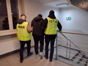 dwóch policjantów w kamizelkach z napisem policja prowadzi zatrzymanego zabójcę korytarzem