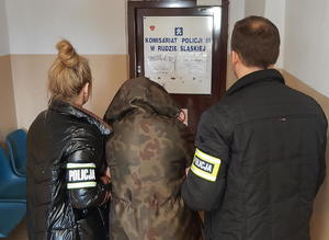 policjantka i policjant po cywilu z opaskami policyjnymi na rękawach prowadzą zatrzymaną korytarzem komisariatu, w tle drzwi z napisem komisariat policji III