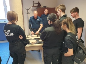 młodzież z klasy policyjnej stoi wokół stolika, na którym leży broń, a policjant pokazuje pistolet maszynowy
