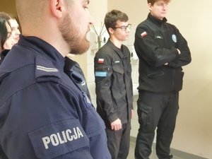 policjant, w tle stoi dwóch uczniów w czarnych mundurach