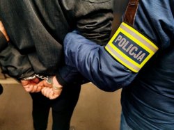 dwie osoby jedna po prawej na rękawie ma napis policja i trzyma drugą za ramie, ręce ma w kajdankach