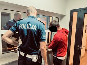 pomieszczenie z szybami, okienko dyżurnego, przed stoi policjant z drogówki, po prawej mężczyzna w czerwonej bluzie z kajdankami na rękach