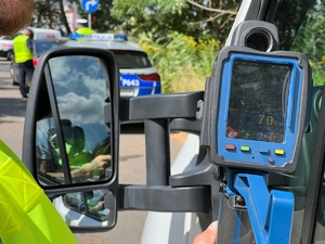 policjant z drogówki przy samochodzie dostawczym pokazuje kierowcy wynik pomiaru ze zdjęciem na którym widać biały samochód i prędkość 70, w tle policjanci i radiowóz