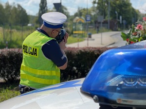 policjant drogówki w trakcie kontroli prędkości, w tle ulica, na pierwszym planie niebieska obudowa lampy błyskowej z radiowozu