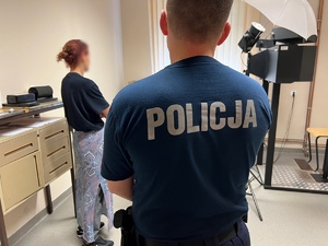 policjant stojący w pomieszczeniu techniki kryminalistycznej gdzie wykonuje się zdjęcia, przed nim stoi kobieta - sprawczyni kradzieży