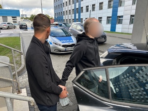 policjant z zatrzymanym przy areszcie w Katowicach, w tle budynki i stojące samochody oraz radiowóz