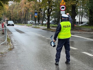 ulica, jadący czarny samochód, policjant z wyciągniętą do góry ręką zatrzymuje kierowcę do kontroli