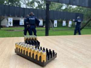 zajęcia strzeleckie na strzelnicy otwartej, na stoliku leży koszyczek z amunicją, w tle strzelający policjanci