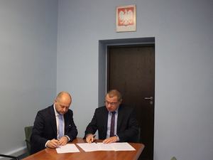 pomieszczenie notariusza, przy stole siedzi Prezydent Miasta i po prawej Komendant Miejski Policji, podpisują dokumenty, na ścianie za nimi godło