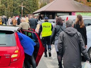 okolica przed cmentarzem, idący ludzie, stojący samochód, pomiędzy widoczny policjant w odblaskowej kamizelce