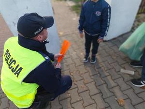 chodnik przed cmentarzem, dzielnicowy kuca przed chłopcem i pokazuje mu opaski odblaskowe