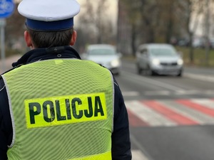 zdjęcie przedstawia policjanta w mundurze stojącego przed przejściem dla pieszych a za przejściem zatrzymują się dwa samochody