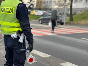 zdjęcie przedstawia policjanta z drogówki stojącego w rejonie przejścia dla pieszych po którym przechodzi kobieta