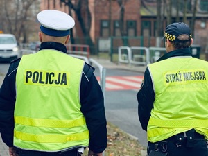 zdjęcie przedstawia policjanta w mundurze i strażnika miejskiego stojących przed przejściem dla pieszych przy szkole