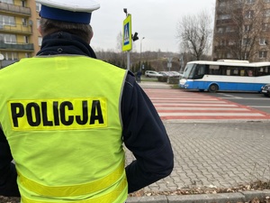 zdjęcie przedstawia policjanta z drogówki stojącego przy przejściu dla pieszych, ulicą jedzie autobus