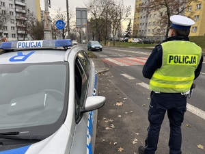 zdjęcie przedstawia policjanta stojącego przy radiowozie i obserwującego drogę i przejście dla pieszych