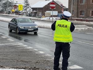 zdjęcie przedstawia policjanta z drogówki na ulicy w trakcie pomiaru prędkości z tyłu widać jadący samochód