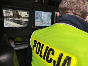 zdjęcie przedstawia policjanta z drogówki w samochodzie monitoringu, w tyle widać ekrany z podglądem z kamer na przejściu dla pieszych