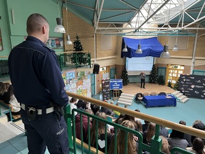 zdjęcie przedstawia, aulę szkolną, stojący policjant w tle uczniowie i kierowca rajdowy