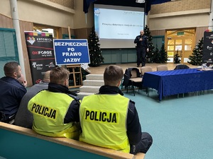 zdjęcie przedstawia, aulę szkolną ze sceną, na której stoi policjant, z przodu na ławce siedzią policjanci