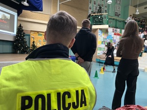 zdjęcie przedstawia siedzącego policjanta i obserwującego jak uczniowie pokonują tor przeszkód w alkogoglach