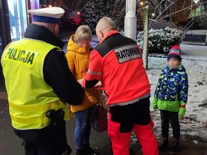 policjant i ratownik przekazuje odblask kobiecie, obok stoi dziecko z opaską odblaskową na ręce