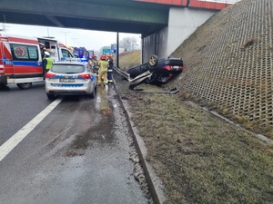 zdjęcie przedstawiające miejsce zdarzenia drogowego, leżący na dachu czarny samochód, na jezdni stoją wozy policji, straży pożarnej i pogotowia
