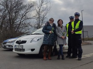Zdjęcie kolorowe przedstawiające policjantów w trakcie kontroli drogowej. Kontrolowana kobieta z otrzymanym od policjantów kwiatkiem