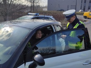 Zdjęcie kolorowe przedstawiające policjanta w trakcie kontroli drogowej. W samochodzie za kierownicą siedzi kobieta z kwiatkiem otrzymanym od policjanta.