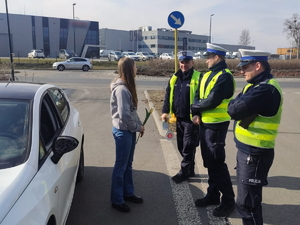 Zdjęcie kolorowe przedstawiające policjantów w trakcie kontroli drogowej. Kontrolowana kobieta z otrzymanym od policjantów kwiatkiem