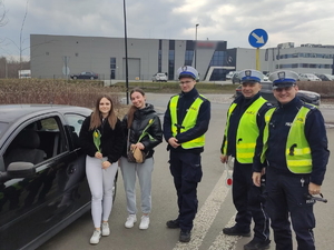 Zdjęcie kolorowe przedstawiające policjantów w trakcie kontroli drogowej, obok samochodu stoją dwie młode kobiety z kwiatkami otrzymanymi od policjantów.