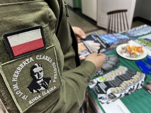 zdjęcie - ramię ucznia w mundurze z naszywkami na rękawie - flaga polski i logo szkoły, w tle stolik z ulotkami wstąp do policji