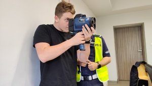 zdjęcie - policjant i chłopak próbujący dokonać pomiaru prędkości ręcznym miernikiem prędkości