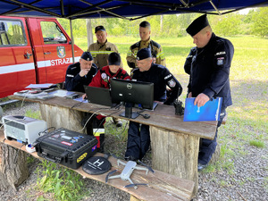 zdjęcie - stolik i ławka ze sprzętem sztabowym, policjanci przyglądają się pracy sztabu poszukiwań