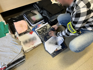 zdjęcie - w pomieszczeniu biurowym rozłożone zarekwirowane ubrania, policjant trzyma kilka bluzek w rękach