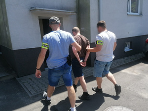 zdjęcie przedstawiające budynek i wejście do niego, w kierunku wejścia idzie dwóch policjantów po cywilu prowadząc zatrzymanego 34-latka