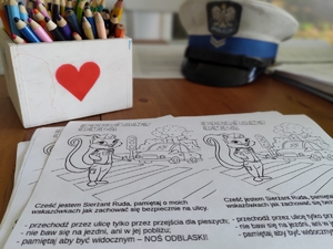 zdjęcie z wizyty policjantów w szkole - czapka policyjna na biurku, kredki, kolorowanka i kartka z sercem