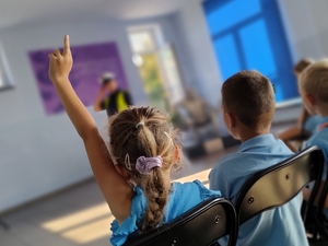 zdjęcie z wizyty policjantów w szkole - dzieci na krzesełkach, dziewczynka z podniesioną ręką, policjant w tle