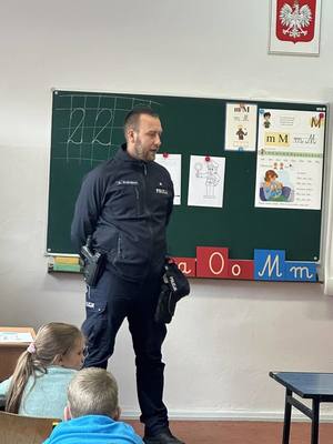 zdjęcie policjanta w trakcie wizyty w szkole, klasa szkolna dzieci w ławkach policjant stoi przy tablicy