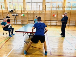 zdjęcie z turnieju przedstawiające salę gimnastyczną, uczniów grających w tenisa stołowego, nauczyciela sędziującego i dzielnicowego, który przygląda się rozgrywkom
